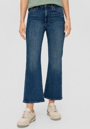 Donkerblauwe flared jeans met een zachte denimtextuur, een midi-taillle, en een ruwe zoom, gecombineerd met lichtgekleurde sneakers.