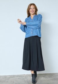 Blusa in denim con scollatura a V e chiusura con bottoni, abbinata a una gonna nera fluida. L'outfit presenta maniche lunghe a sbuffo e stivali alla caviglia.