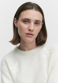 Jonge vrouw met stijl bruin haar, blauwe ogen, die kleine oorbellen in de vorm van een ring draagt en een witte trui met textuur en bloemdetails.