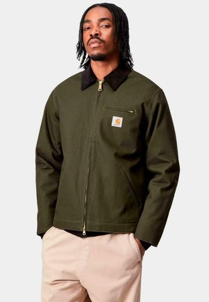 Uomo con capelli intrecciati, che indossa una giacca verde oliva con zip e colletto nero e pantaloni beige, in piedi con le mani in tasca.
