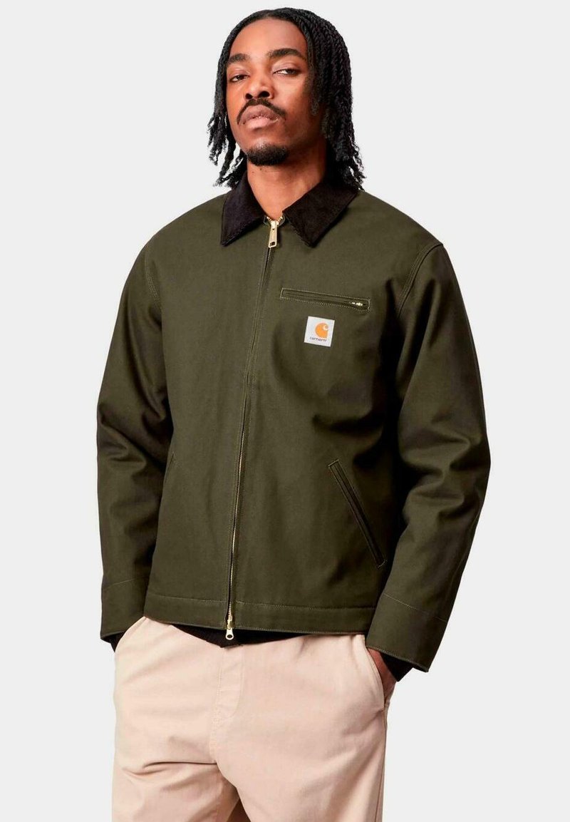 Uomo con capelli intrecciati, che indossa una giacca verde oliva con zip e colletto nero e pantaloni beige, in piedi con le mani in tasca.