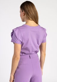 Paarse korte mouw T-shirt met opstaande schouders, een gladde textuur, ronde halslijn, gezien vanuit de achterkant. Gepaard met strakke paarse broek.