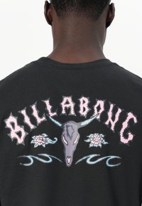Svart bomull T-skjorte med grafisk design av et hodeskalle, blomster, og teksten "BILLABONG" i rosa og blå nyanser.