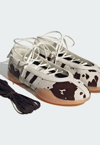 Par lavtoppede joggesko med hvitt og brunt ku-skinnmønster, beige lisser, gummisåler og et ekstra sett med svarte lisser ved siden av.