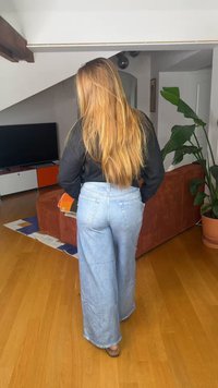Frau mit langen blonden Haaren, die ein schwarzes Oberteil und weite blaue Jeans trägt, steht in einem Wohnzimmer mit braunem Sofa und grüner Pflanze.