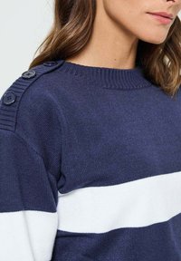 Pull en maille bleu marine avec une rayure horizontale blanche, doté d'un col et d'un détail d'épaule côtelés et de quatre boutons foncés.