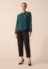 Teal blouse with long, sheer sleeves, gathered neckline, and tie detail; paired with black tapered trousers and black heels.