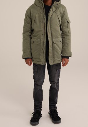 Personne portant une parka à capuche vert olive, un pantalon cargo noir et des baskets noires, debout devant un fond beige uni.