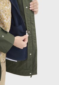 Veste imperméable verte avec une fermeture éclair, dévoilant une doublure en polaire marine et une chemise intérieure à motif floral. Comprend des boutons pression et des poches.