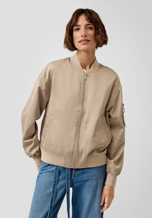 Frau mit kurzem braunem Haar, trägt eine beigen Bomberjacke und blaue Jeans, steht vor einem schlichten hellgrauen Hintergrund.