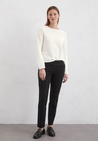 Weißer langärmeliger Pullover kombiniert mit schwarzen Slim-Fit-Hosen und schwarzen Slippern. Glattes Material, einfaches Design und neutrale Farben.