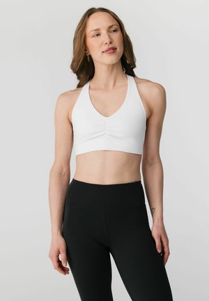 Világos bőrű, barna hajú nő fehér, pánt nélküli sportmelltartót és fekete, magas derekú leggingset visel, egy egyszínű világos háttér előtt áll.