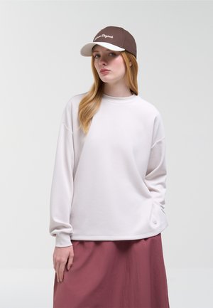 Jeune femme aux longs cheveux blonds portant un sweat blanc, une jupe mauve et une casquette de baseball marron et blanche, sur un fond uni.