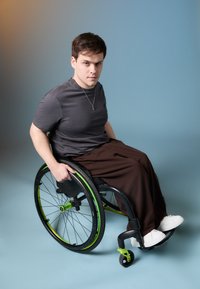 Αμαξίδιο με μαύρο σκελετό και πράσινες λεπτομέρειες, κατειλημμένο από ένα άτομο με γκρι μπλούζα και καφέ παντελόνι, με φόντο μπλε.