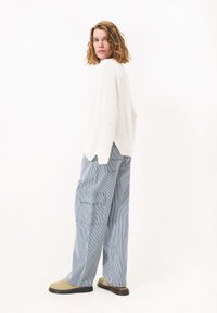 Witte gebreide trui met een losse pasvorm, gecombineerd met blauw en wit gestreepte cargobroek. Comfortabele beige instappers met minimale detailing.