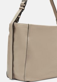 Beżowa skórzana torebka o gładkiej fakturze, z minimalistycznym designem, regulowanym paskiem i subtelnym czarnym przeszyciem wzdłuż krawędzi.