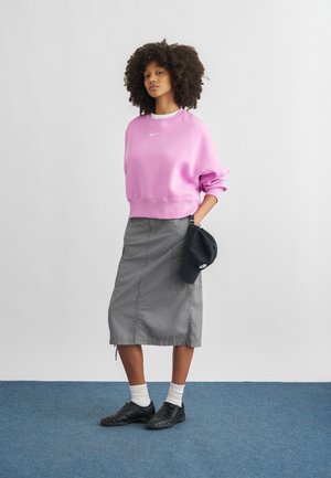 Mujer con cabello rizado que lleva una sudadera rosa de Nike, falda gris hasta la rodilla, calcetines blancos, zapatos negros y sostiene una gorra negra de Nike a un lado.