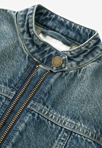 Farmerdzseki magas gallérral, sötétkék cipzárral és réz hardverelemekkel. Jellemzője a texturált anyag, látható varrásokkal és fém gombbal.