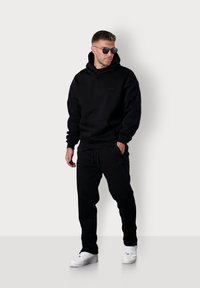 Black fleece hoodie with kangaroo pocket, paired with black sweatpants. Worn with white sneakers and round sunglasses. Minimal branding visible.