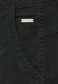 Černá tkanina s hladkým povrchem, s viditelným šitím a stříbrnou štítkem s vyrytým nápisem "JACK & JONES" připojeným k kapse.