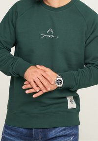 Uomo che indossa una felpa Jack & Jones verde scuro e un orologio d'argento con quadrante nero, mani incrociate davanti, visibili jeans blu.