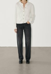 Valkoinen neulecardigan, jossa on kultaiset napit ja taskut, yhdistettynä tummiin suoriin farkkuihin ja mustiin loafer-kengiin, neutraalilla taustalla.