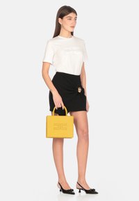 Bolso de mano de cuero amarillo con forma estructurada, con logo grabado y detalles cosidos, sostenido por una modelo que lleva una camiseta blanca y una falda negra.