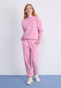 Sweat-shirt rose et jogging assorti en tissu doux, avec un logo sur la poitrine et sur le côté. Le mannequin se tient sur un sol bleu devant un fond uni.