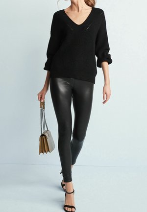 Schwarzer Strickpullover mit V-Ausschnitt, kombiniert mit glänzenden schwarzen Leggings. Eine kleine graue Handtasche mit Kettenakzenten vervollständigt das Outfit.