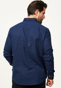 Dunkelblaues Langarmhemd mit spitzem Kragen, geknöpften Manschetten, einer dezenten Textur und einem abgerundeten Saum, von hinten betrachtet.