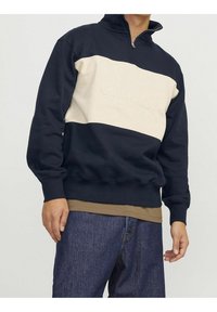Hombre vistiendo sudadera de media cremallera en azul marino y beige con el texto "ORIGINALS", superpuesta sobre una camisa marrón, combinada con jeans azules.
