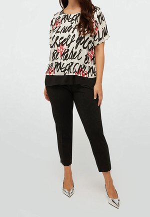 Mujer con camiseta blanca de manga corta con texto en negro y flores rojas, pantalones negros cortos y tacones plateados puntiagudos, de pie sobre fondo liso.