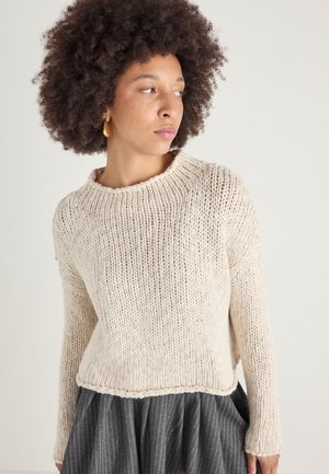 Pull en maille texturée crème avec une coupe décontractée, un large col rond et des poignets côtelés, associé à une jupe grise à rayures.