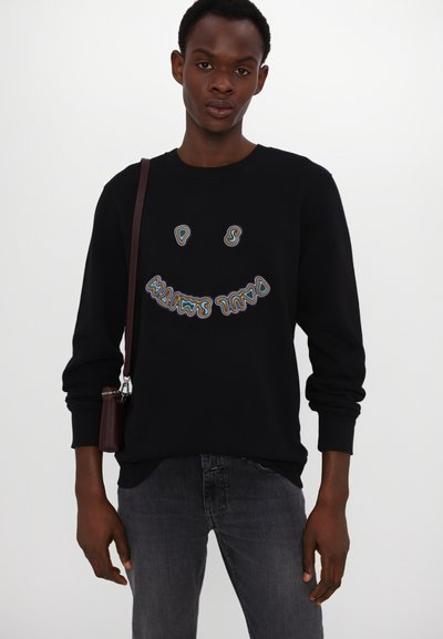 Sudadera negra con cuello redondo, mangas largas y un diseño de cara sonriente bordado y colorido. La modelo lleva unos jeans gris oscuro y sostiene un bolso pequeño.