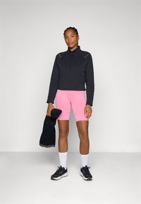 Sweat-shirt noir zippé avec des accents réfléchissants, associé à un short de cyclisme rose. Le modèle tient une serviette noire, portant des baskets noires et des chaussettes blanches.