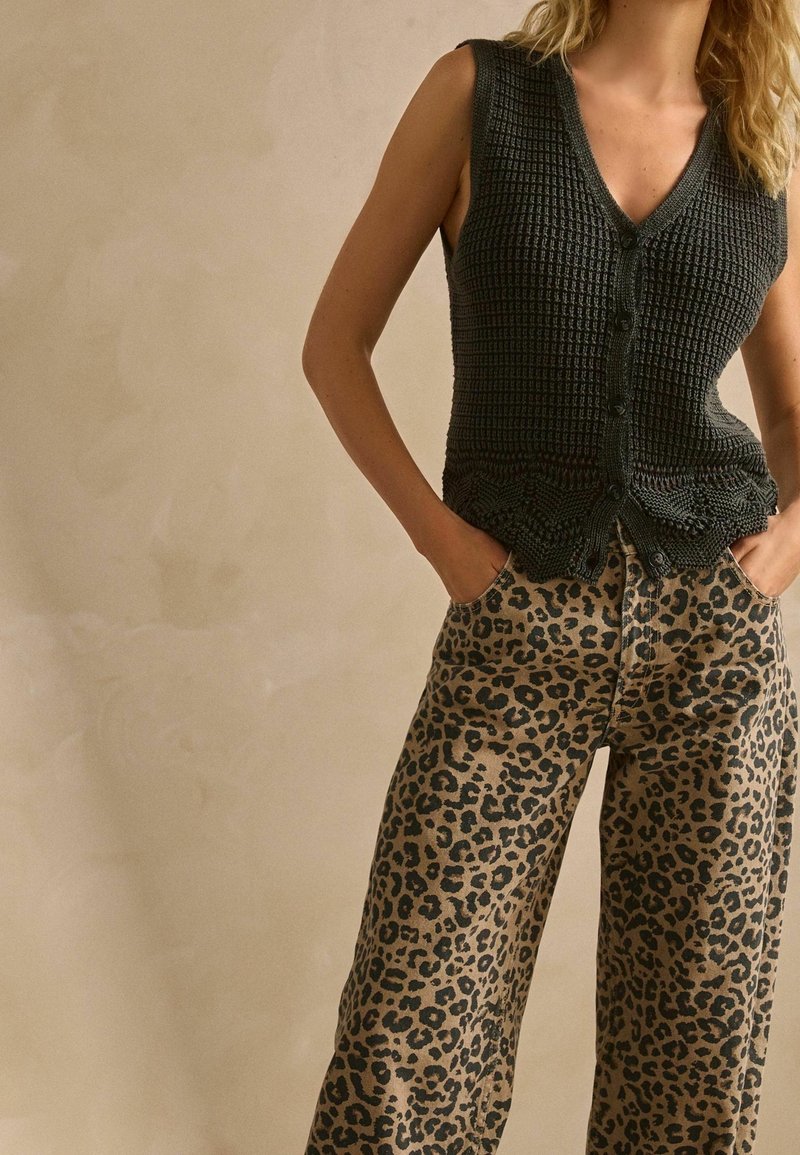 Femme portant un gilet noir en maille sans manches boutonné et un pantalon taille haute imprimé léopard, les mains dans les poches, sur un fond uni.
