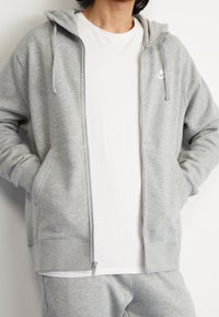 Persona che indossa una felpa Nike grigia chiara con zip sopra una maglietta bianca e pantaloni della tuta grigi abbinati, con le mani in tasca.