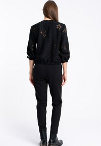 Kobieta w czarnej, półprzezroczystej bluzce w wzory z rękawami 3/4, czarnych dopasowanych spodniach i czarnych botkach za kostkę, odwrócona plecami na białym tle.