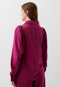 Camicia lunga a maniche viola con colletto, maniche a sbuffo e yoke posteriore a forma di V. Texture morbida e vestibilità leggermente ampia.