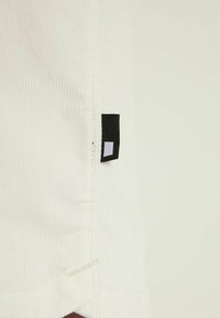 Nahaufnahme von weißem geripptem Stoff mit einem kleinen schwarz-weißen rechteckigen Etikett, das nahe der Naht genäht ist.