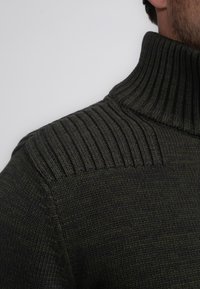 Mörkgrön stickad tröja med hög ribbad krage och texturerad axeldetalj, med en slät kropp och figurnära silhuett.