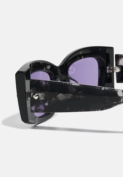 Occhiali da sole neri di grandi dimensioni con lenti viola. La montatura presenta un design robusto e braccia scure testurizzate, fissate con dettagli metallici.