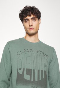 Sudadera verde con cuello redondo, que presenta un texto en negrita de color gris oscuro: "RECLAMA TU DENIM CONSTRUIDO EN AZUL" en el frente. Tejido de algodón suave.
