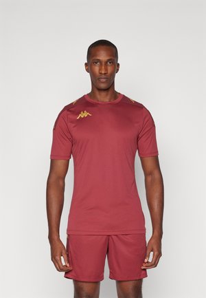 Muž oblečený v bordovom športovom oblečení so zlatými logami na ramenách a hrudi, stojaci pred jednofarebným svetlým pozadím.