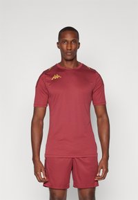 Bordeaux sportshirt en shorts, gemaakt van gladde stof. Heeft gouden accenten op de schouders en het logo, met een ronde hals en korte mouwen.