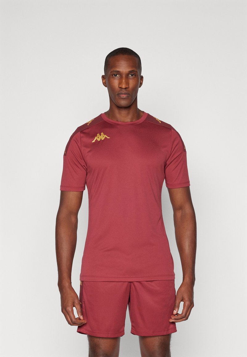 Bordeaux sportshirt en shorts, gemaakt van gladde stof. Heeft gouden accenten op de schouders en het logo, met een ronde hals en korte mouwen.
