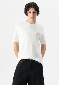 Witte katoenen T-shirt met korte mouwen, ronde hals en een kleine rode grafische print op de linkerborst. Gemaakt met donkere denim jeans.