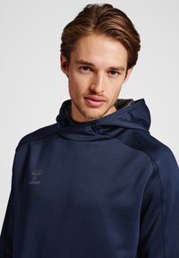 Granatowa bluza z kapturem o miękkiej fakturze, z wysokim kołnierzem i logo na piersi. Minimalistyczny design z dopasowanym krojem.