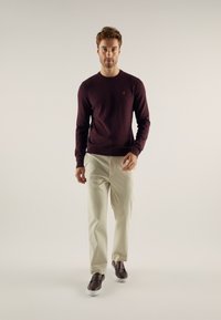Bordo pulover, krema hlače i smešani cipele. Pullover ima mali logo na grudima. Model stoji protiv neutralne pozadine.