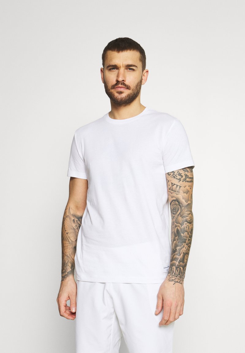 White cotton t-shirt with a round neck and short sleeves. Smooth texture, classic fit, displayed on a person with tattoos on the forearm.